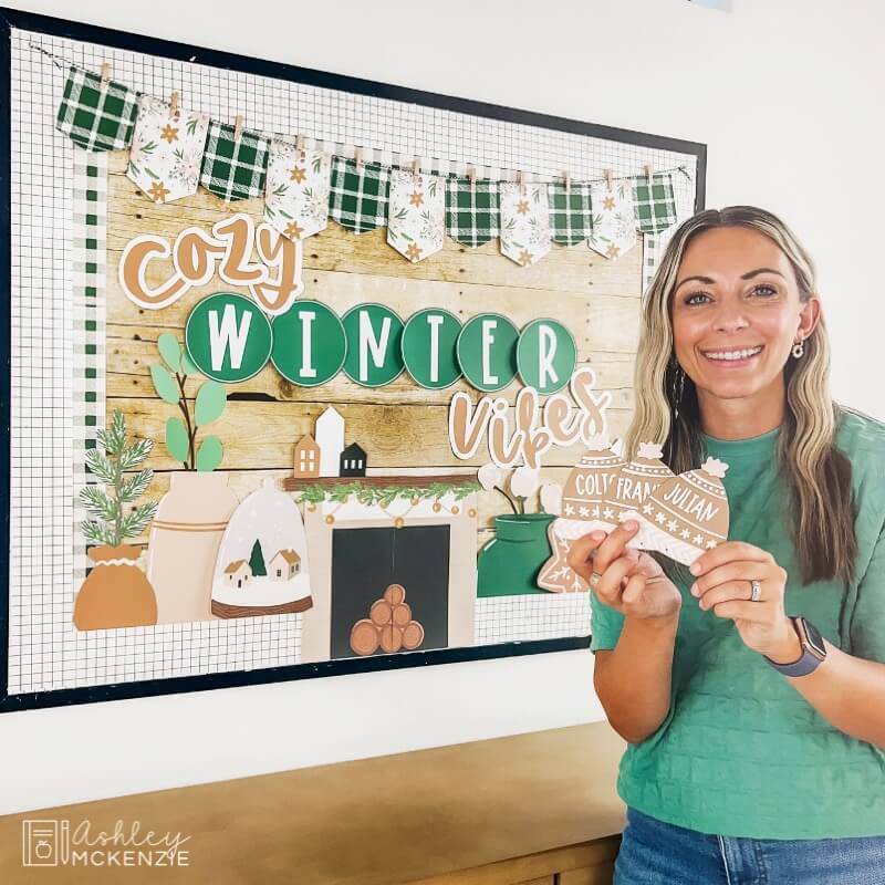 A classroom bulletin board features the saying "Cozy Winter Vibes" with gold and green tones, including a fireplace, winter greenery, and winter hats with students' names written on them to be displayed on the board.