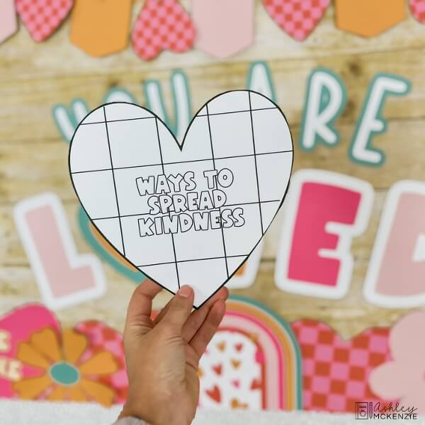 A heart template is held up with the words "Ways to Spread Kindness" on it, to compliment Valentine's Day Classroom Decor.
