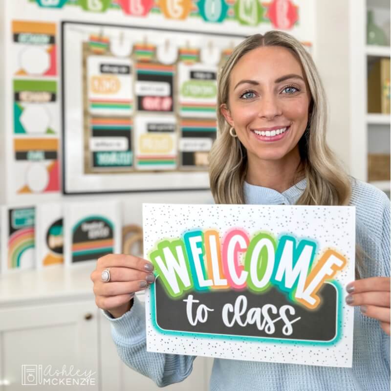 A teacher holding up a brightly colored sign that says "Welcome to Class"