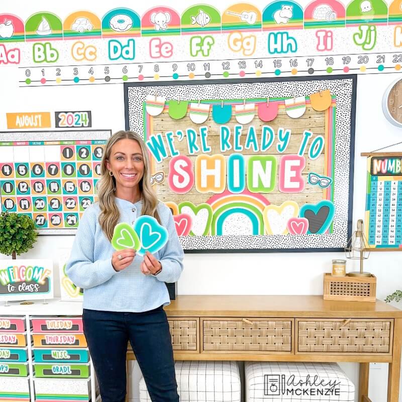 A teacher standing in front of a bulletin board decorated in a bright neon classroom decor theme with the saying "We're ready to shine"
