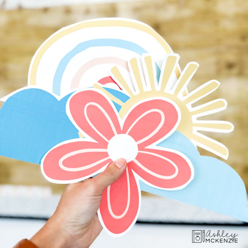 Spring classroom decor cutouts are held up, including a sun, pink flower, cloud, and rainbow