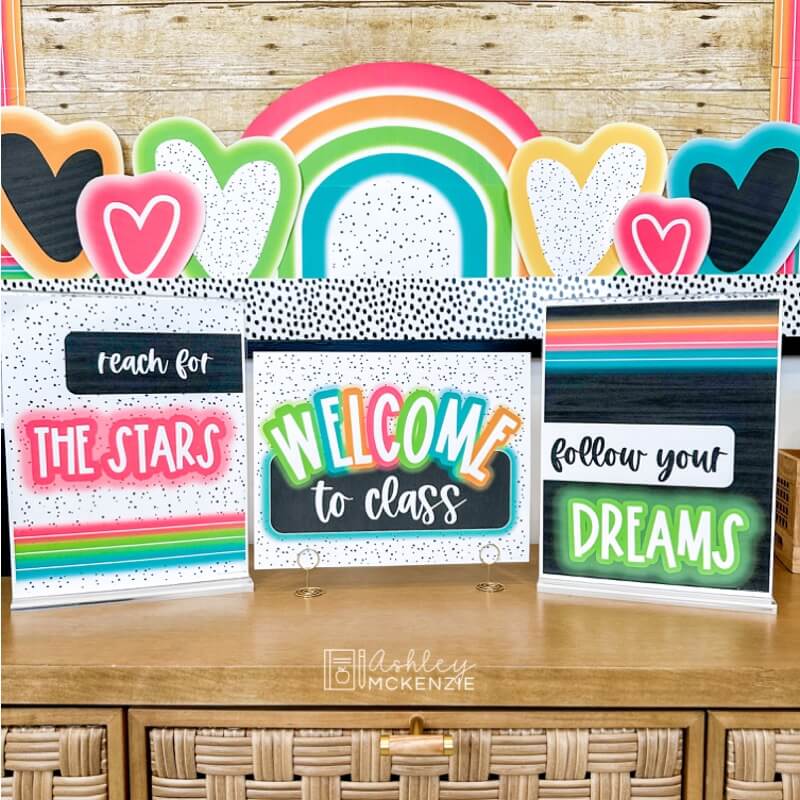 Three brightly colored posters are displayed on a classroom table with the sayings "Welcome to Class", "Reach for the stars," and "follow your dreams"